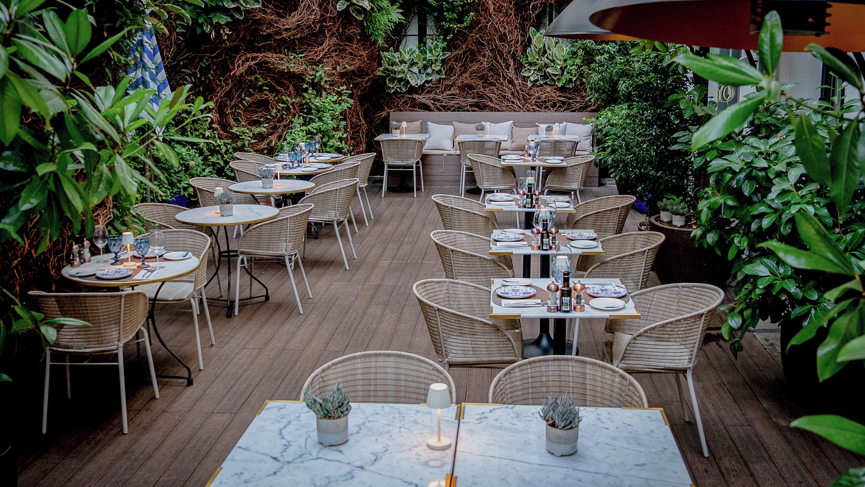 An outdoor patio with wicker furniture and marble-topped tables, surrounded by lush greenery.