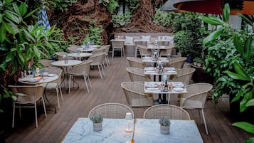 An outdoor patio with wicker furniture and marble-topped tables, surrounded by lush greenery.
