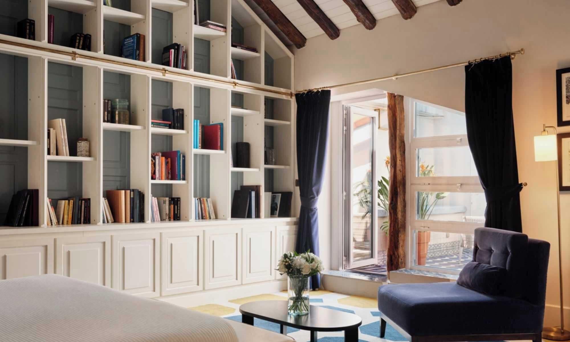 A hotel room with a large bookshelf, a comfortable sofa, a small round table, and a view through a window with curtains.