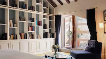 Ein Hotelzimmer mit einer großen Bücherwand, einem gemütlichen Sofa, einem kleinen runden Tisch und Blick durch ein Fenster mit Vorhängen.