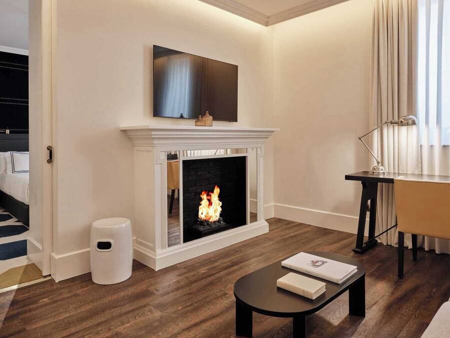 A modern living room with a fireplace, a flat-screen TV above it, a desk with a lamp, and a sofa.