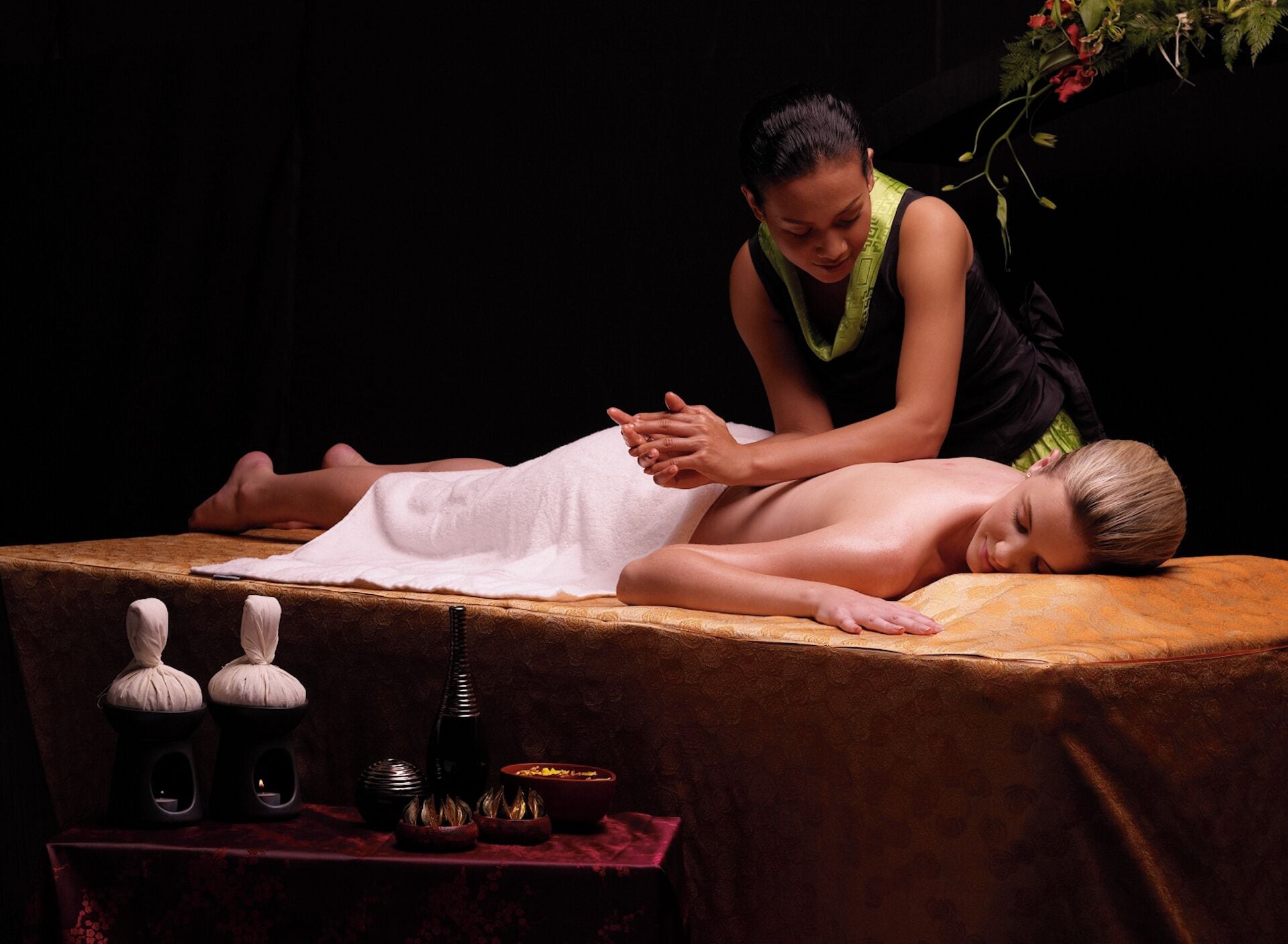 A massage therapy session with a person lying face down on a massage table, another person attending to them, and various massage tools on a table.