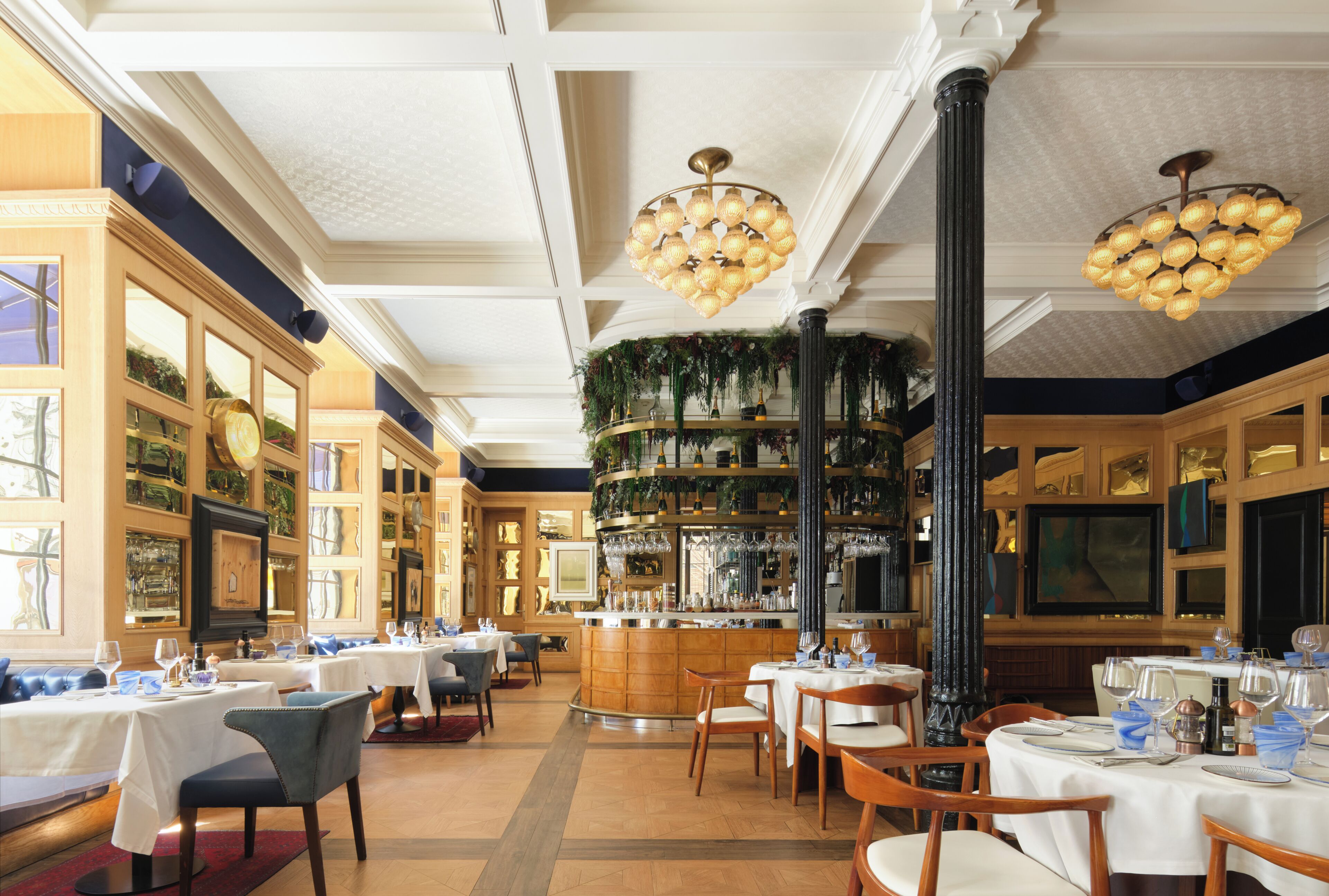 A restaurant interior with multiple tables, chairs, a bar area, and decorative plants.