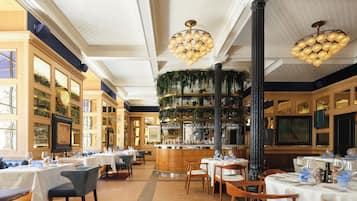 A restaurant interior with multiple tables, chairs, a bar area, and decorative plants.