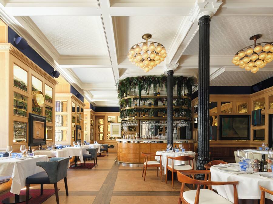 A restaurant interior with multiple tables, chairs, a bar area, and decorative plants.