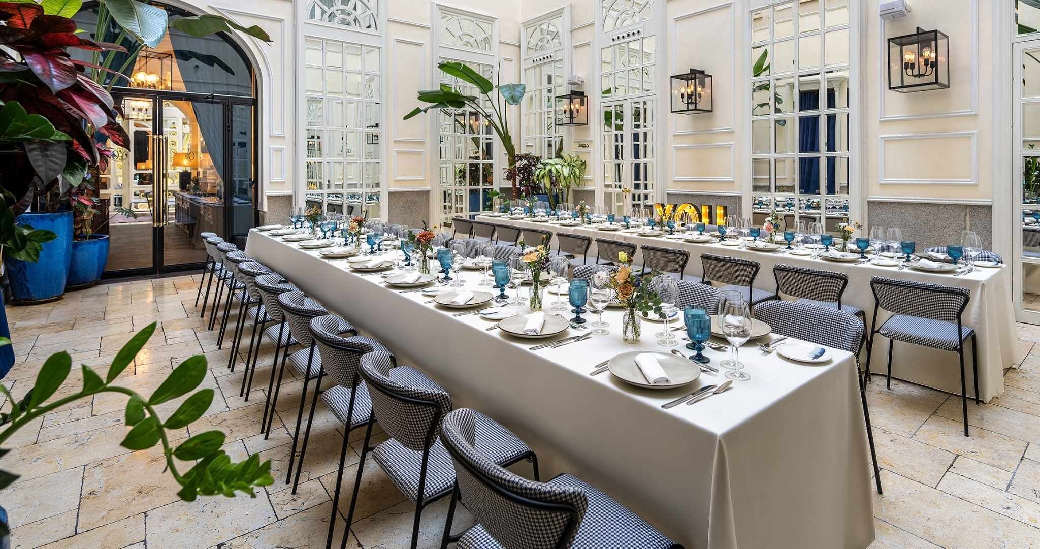 Una larga mesa de comedor dispuesta para una comida en un ambiente bien iluminado con ventanas amplias y plantas en macetas.