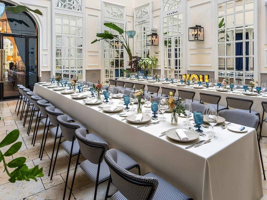 A long dining table set for a meal in a well-lit room with large windows and potted plants.