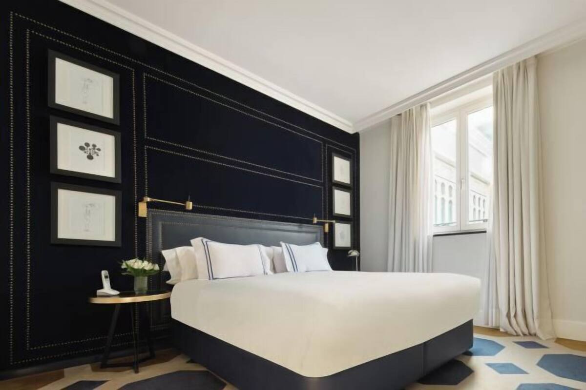 Une chambre d’hôtel avec un grand lit, une table de chevet avec une lampe, des tableaux encadrés au mur et une fenêtre avec des rideaux.
