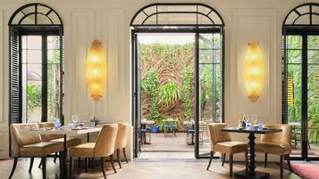 A dining area with tables and chairs, large windows, and a view of greenery outside.