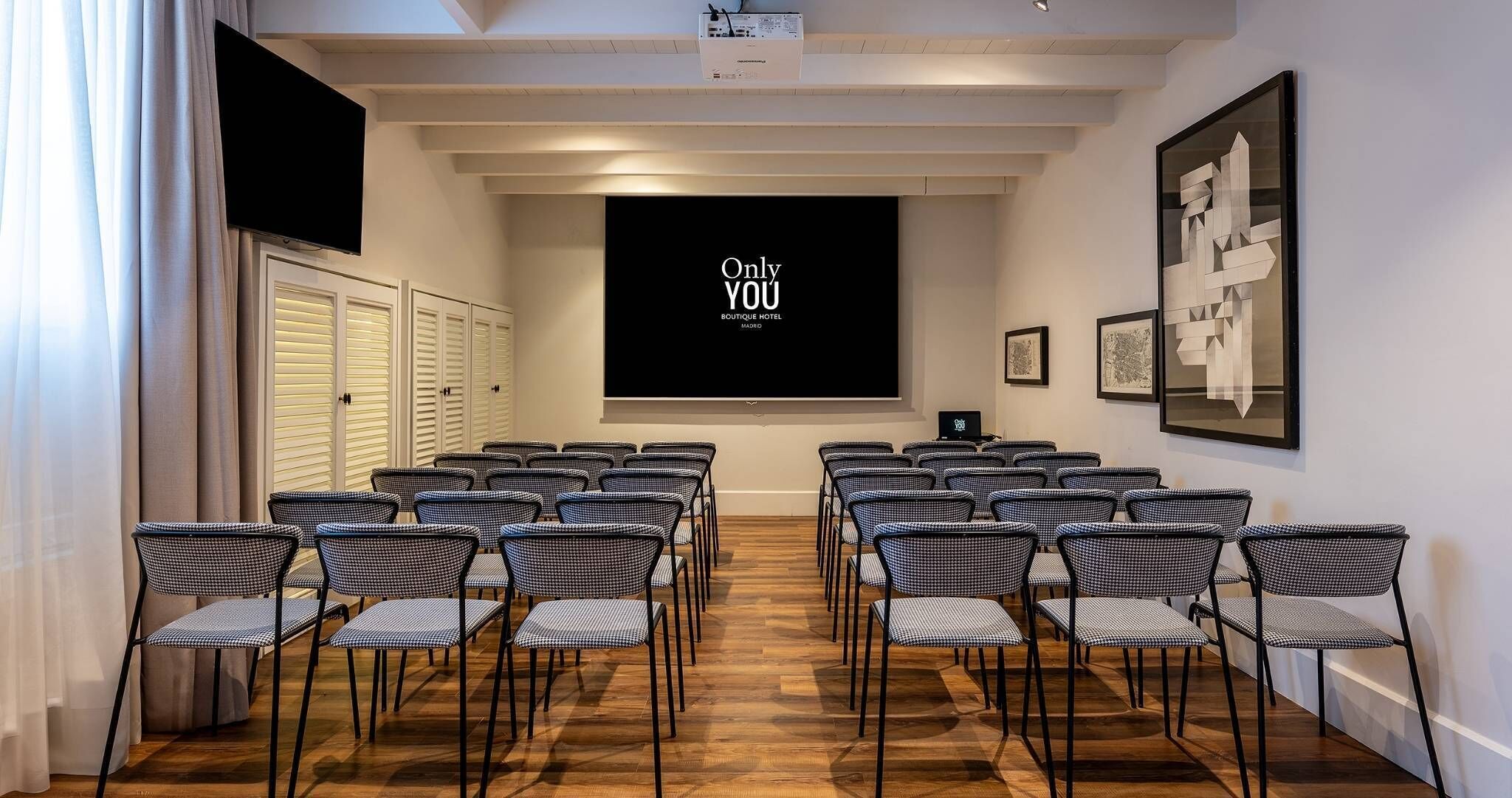 A conference room with rows of checkered chairs, a projector screen displaying 'Only YOU BOUTIQUE HOTEL Madrid', and framed artwork on the walls.