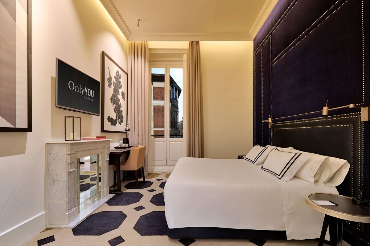 A hotel room with a bed, a desk, a chair, a TV, and a view of the city.