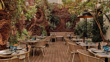 An outdoor dining area with wicker tables and chairs, surrounded by lush greenery and vines.