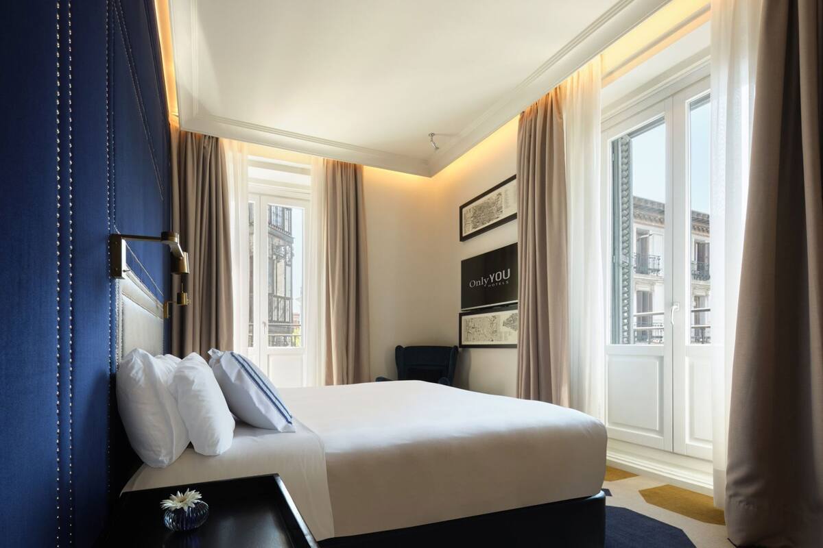 Une chambre d’hôtel avec un lit, une table de chevet, un téléviseur fixé au mur et une fenêtre avec des rideaux.