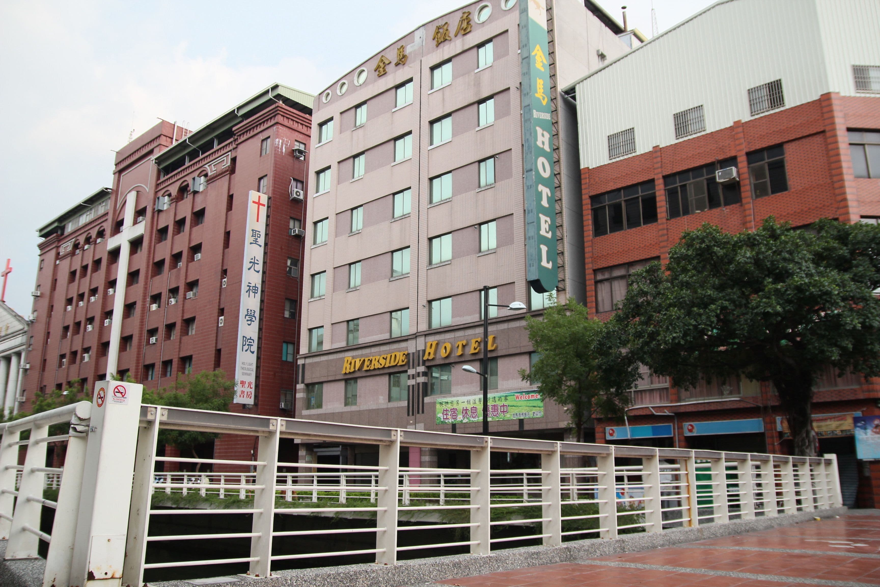 A multi-story hotel with a prominent Riverside Hotel sign, featuring a traditional architectural style with brickwork and multiple windows.
