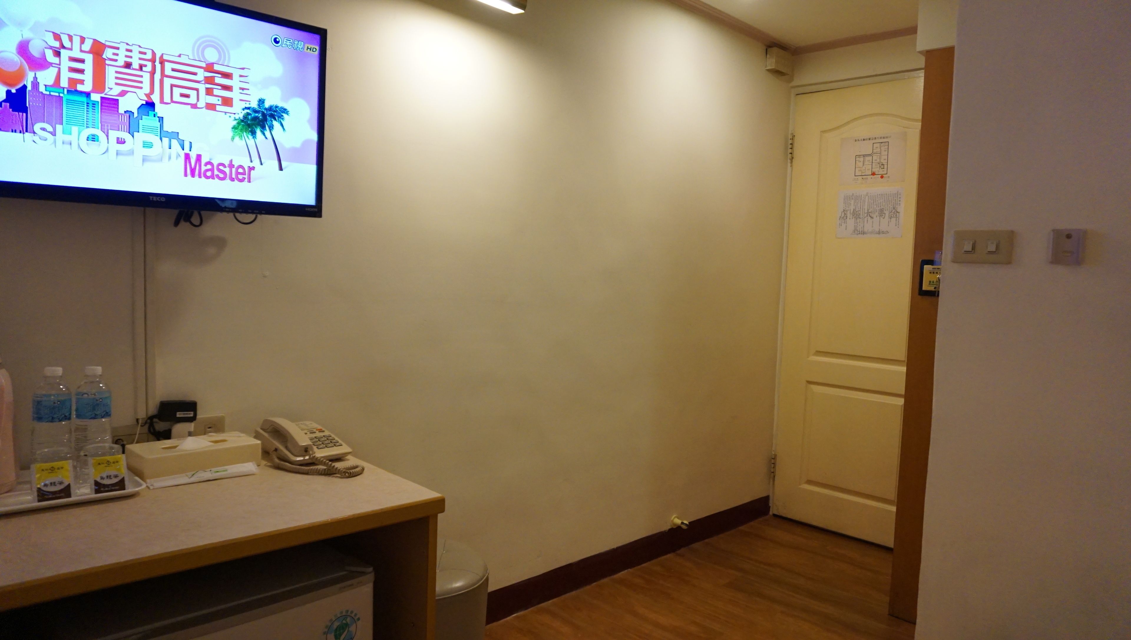 A hotel room with a desk, a telephone, bottled water, and a television displaying a shopping channel.