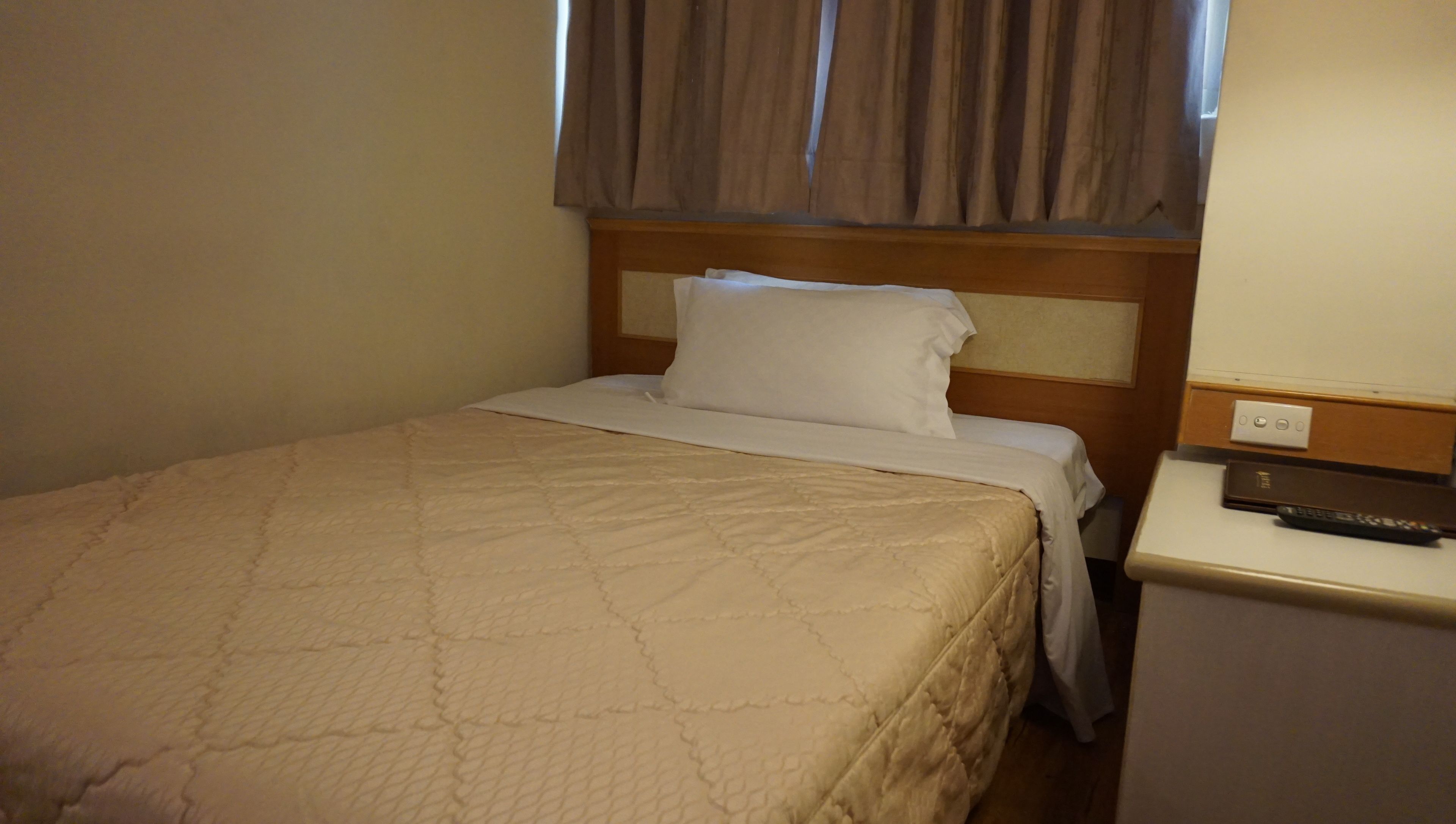 Een enkel bed met een gewatteerde sprei, een houten hoofdbord, een wit kussen, een nachtkastje met een afstandsbediening en een stopcontact.