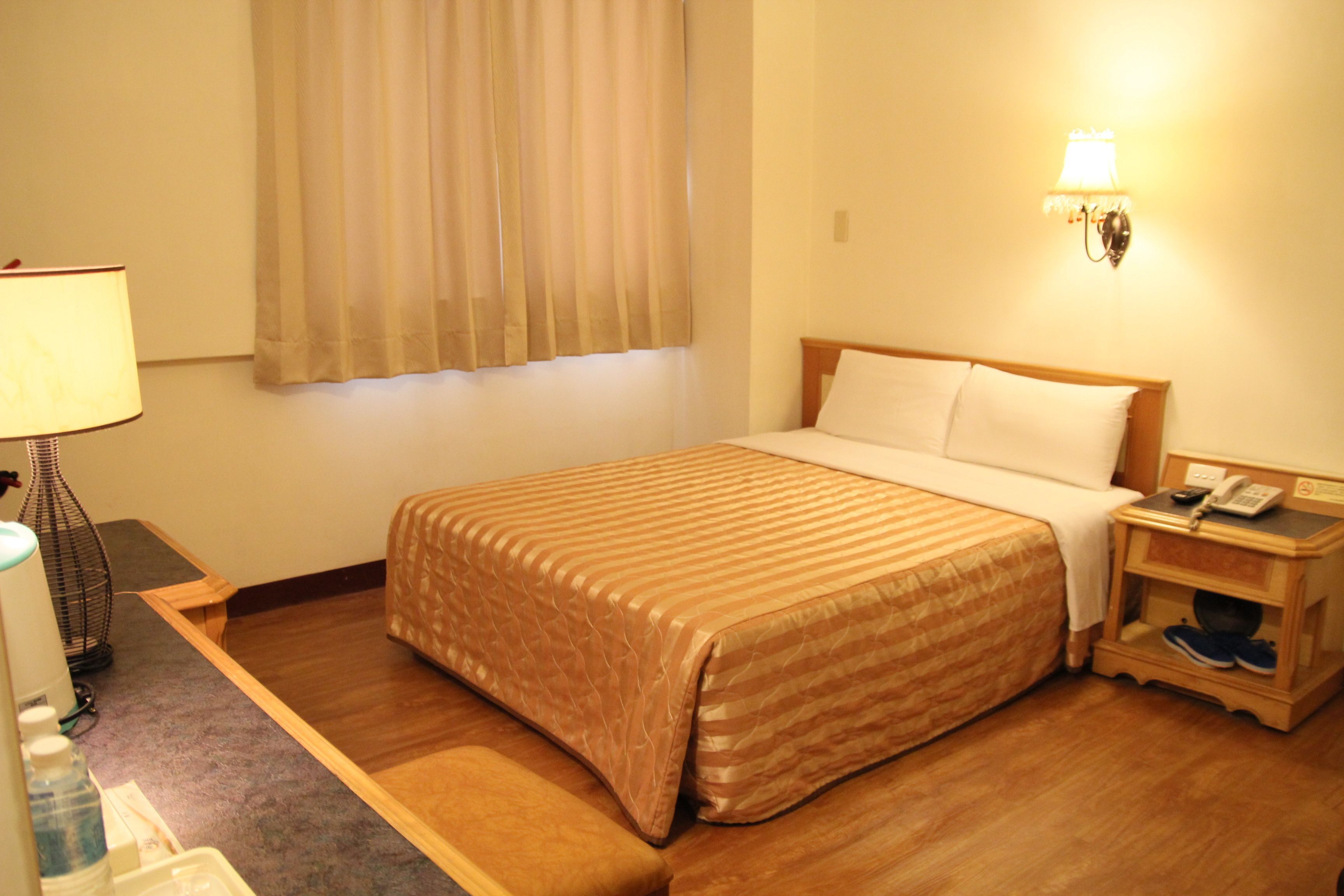 A hotel room with a bed, bedside table, lamp, and a window with curtains.