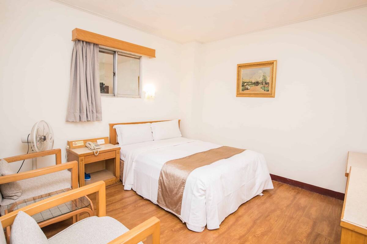Ein Hotelzimmer mit Bett, Schreibtisch, Stuhl, Ventilator und einem Bild an der Wand.