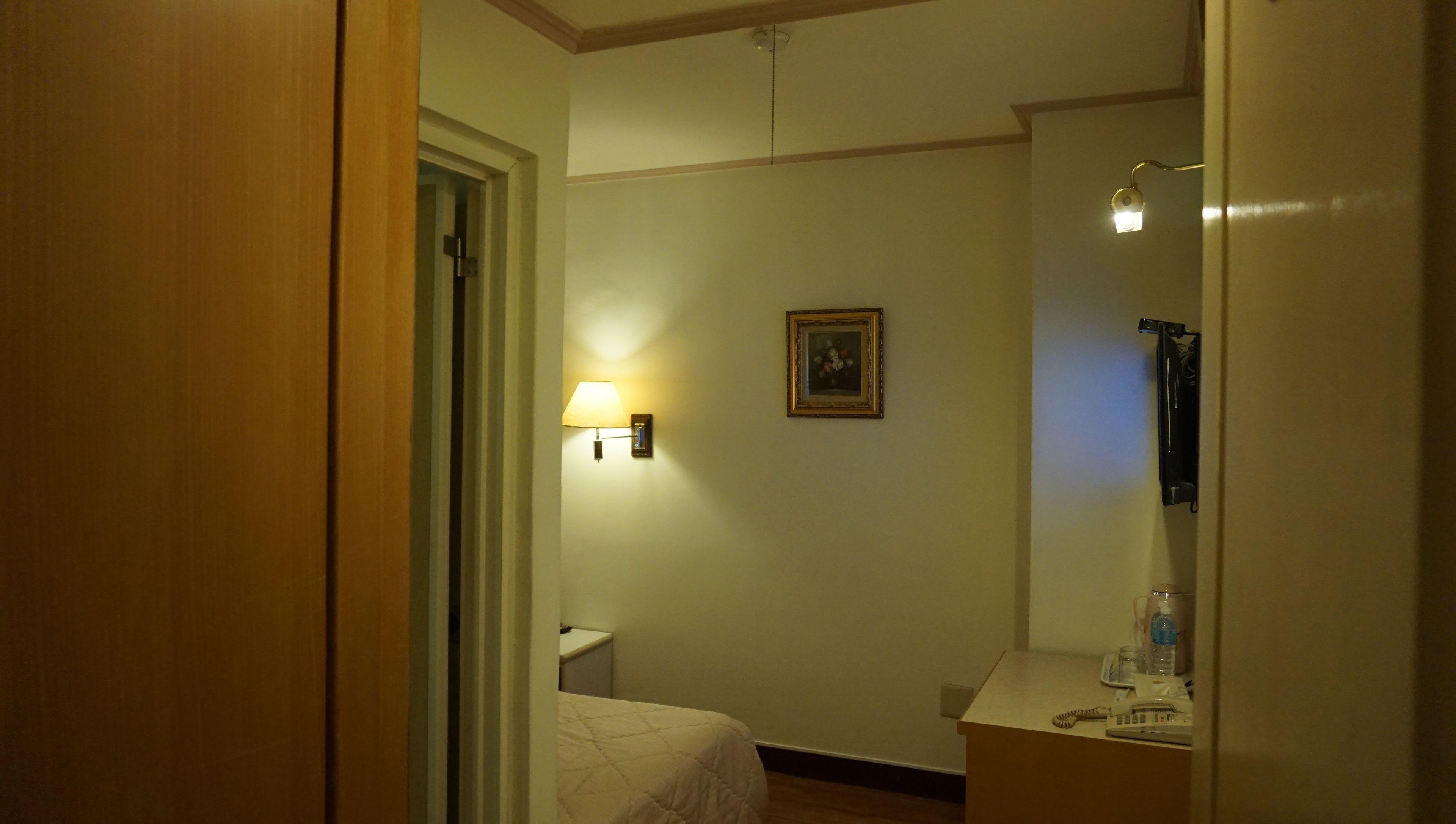 Ett hotellrum med en säng, ett nattduksbord med en lampa, en inramad tavla på väggen och en spegel.