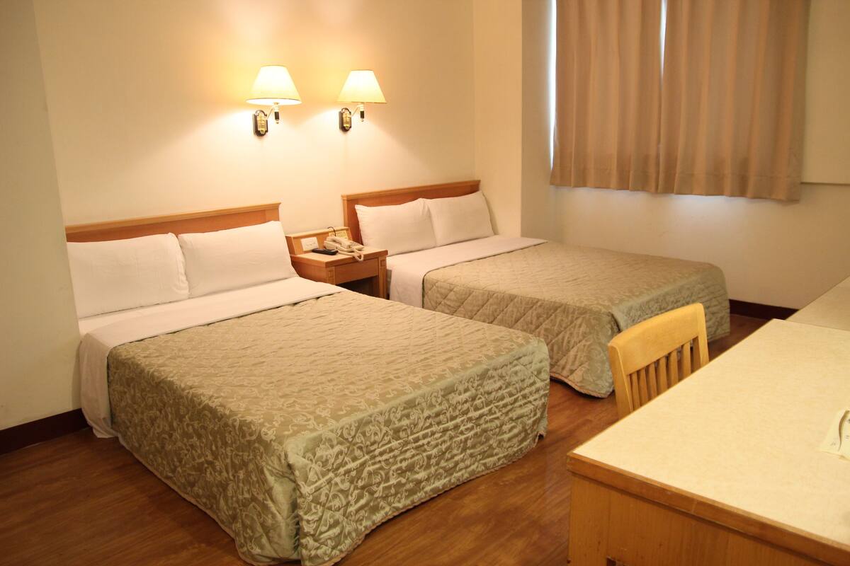 Ein Hotelzimmer mit zwei Betten, einem Nachttisch aus Holz mit Telefon, einem Tisch und zwei Wandlampen.