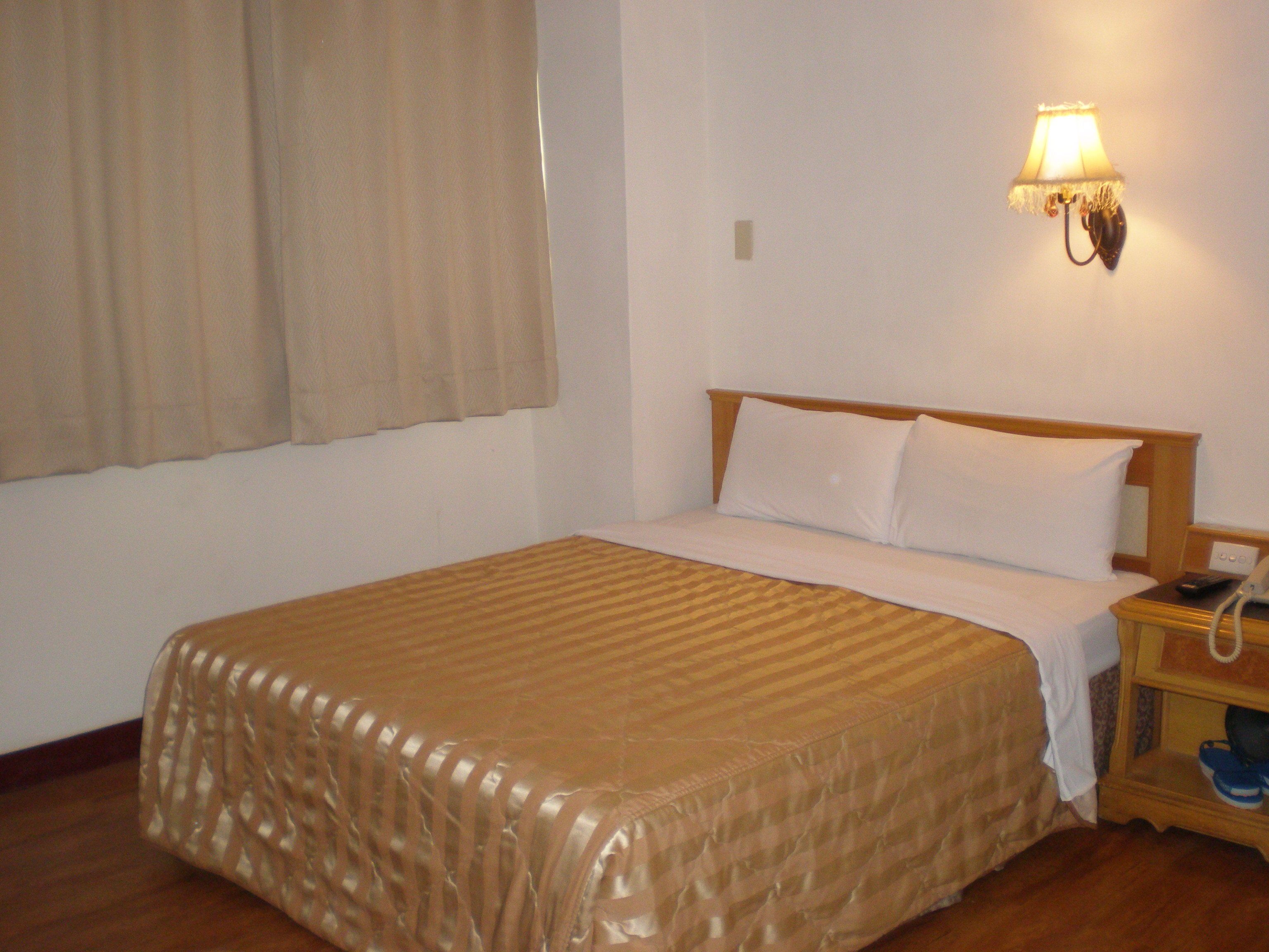 A hotel room with a bed, a nightstand with a phone and slippers, and a wall-mounted lamp.