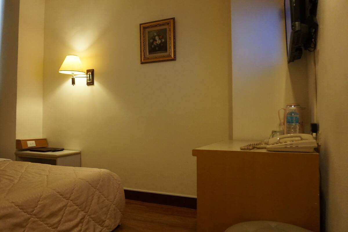 Ein Hotelzimmer mit Bett, Nachttisch, Lampe, einem Bild an der Wand und einem Schreibtisch mit Telefon und Wasserflasche.