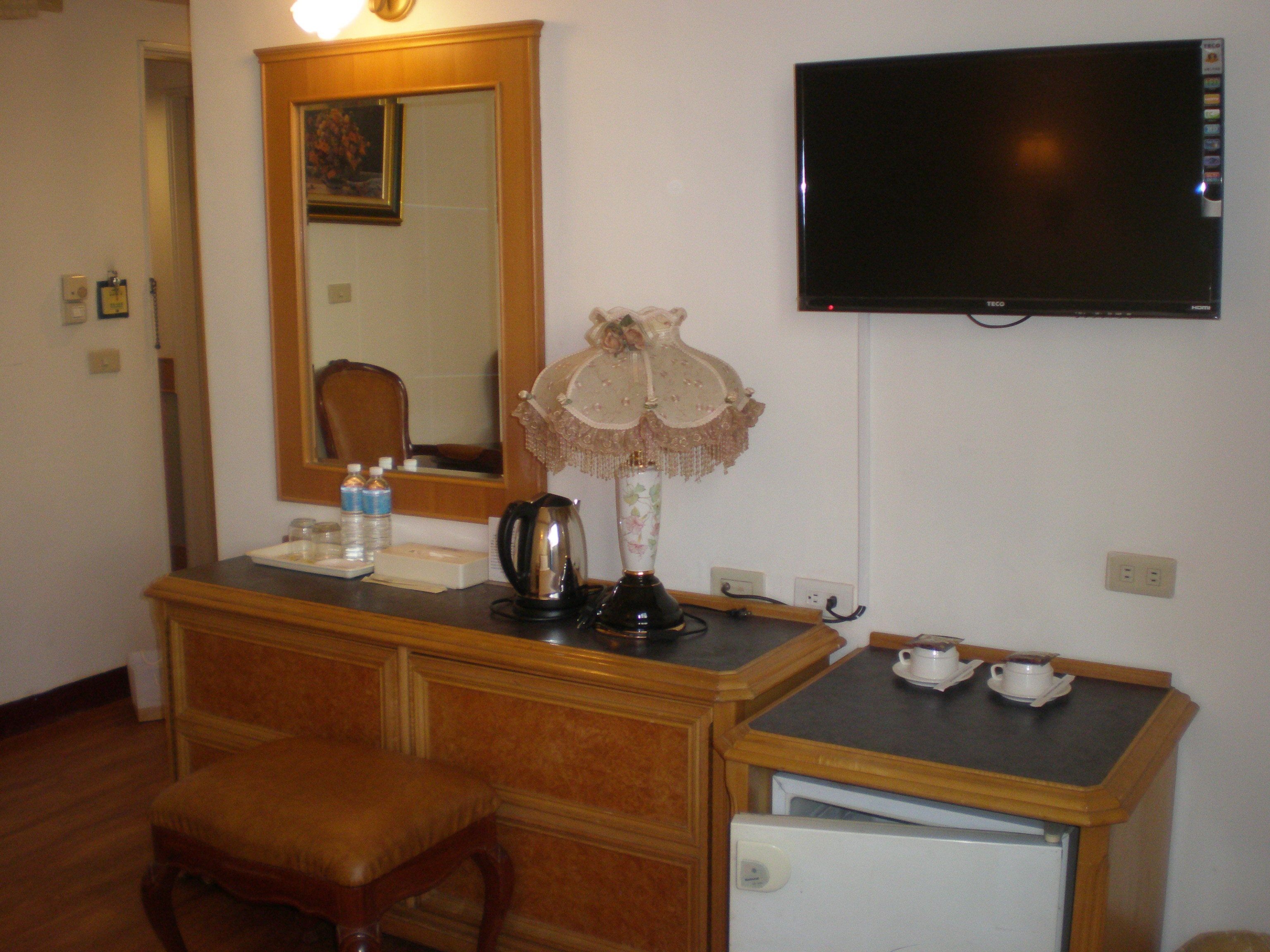 A room with a wooden dresser, a small table with a kettle and cups, a television, and a mirror.