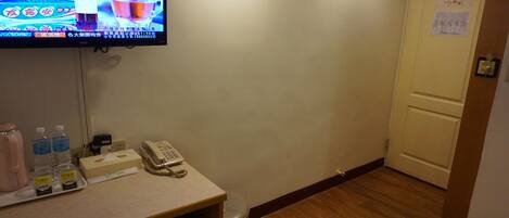 A hotel room with a television displaying an advertisement, a telephone, a trash bin, and a small table with bottled water and a tissue box.