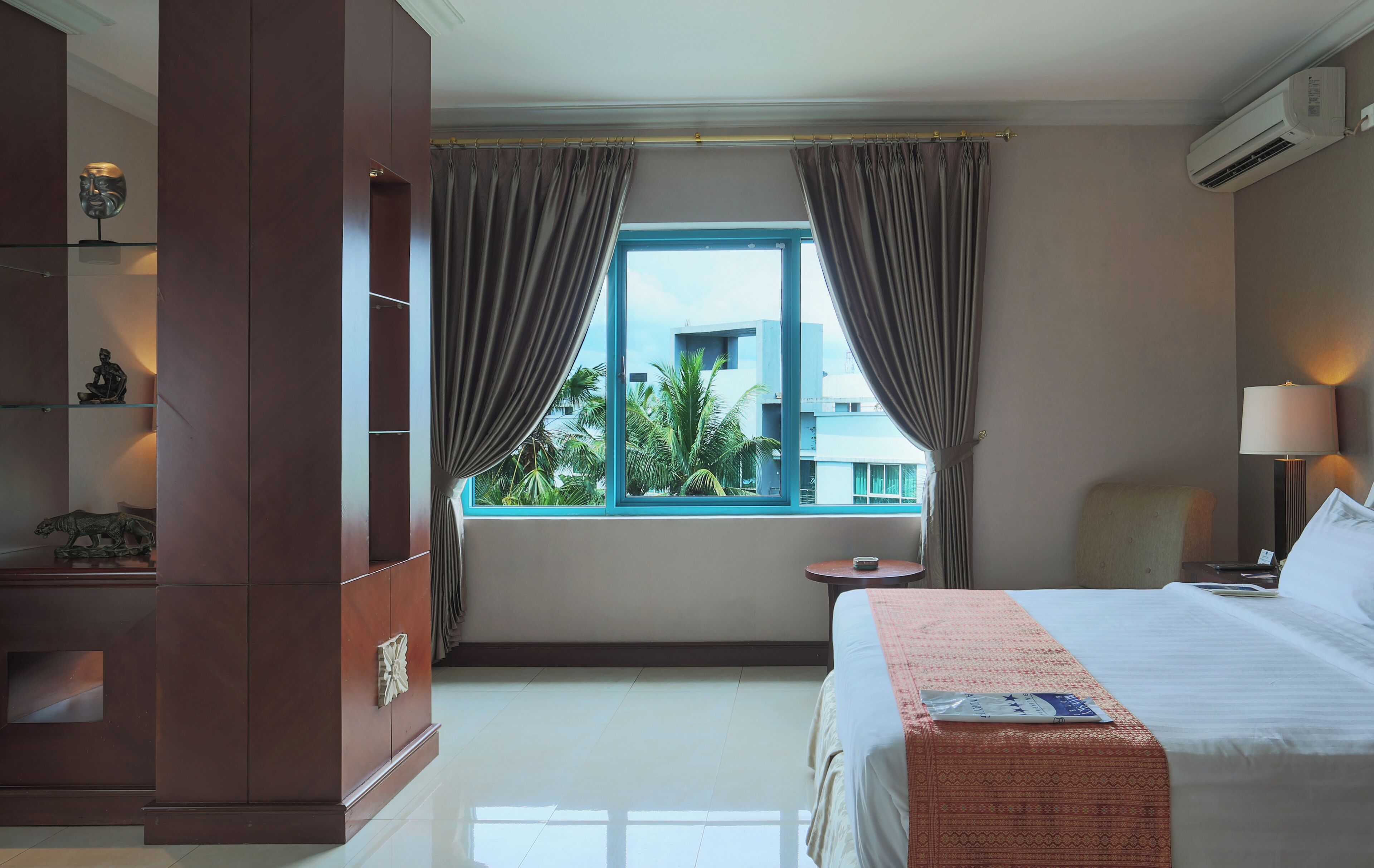 Habitación de hotel con una cama, una mesita de noche, una lámpara, una ventana con cortinas y vistas a edificios.