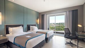 A hotel room with two beds, a desk, and a chair. There is a balcony with a view of the city. - Blue Sky Hotel (Balikpapan)