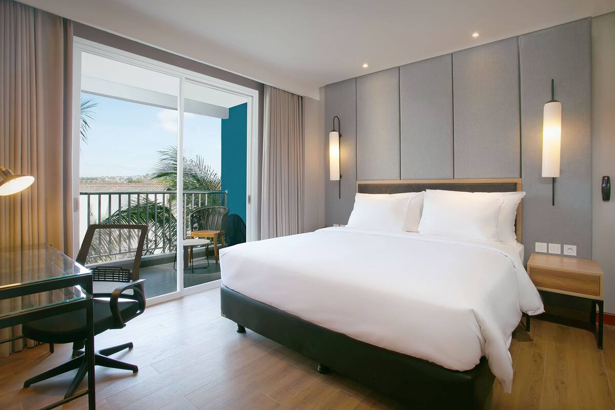 Una habitación de hotel con una cama grande, un escritorio con una silla, un balcón con vistas y una lámpara de pared.