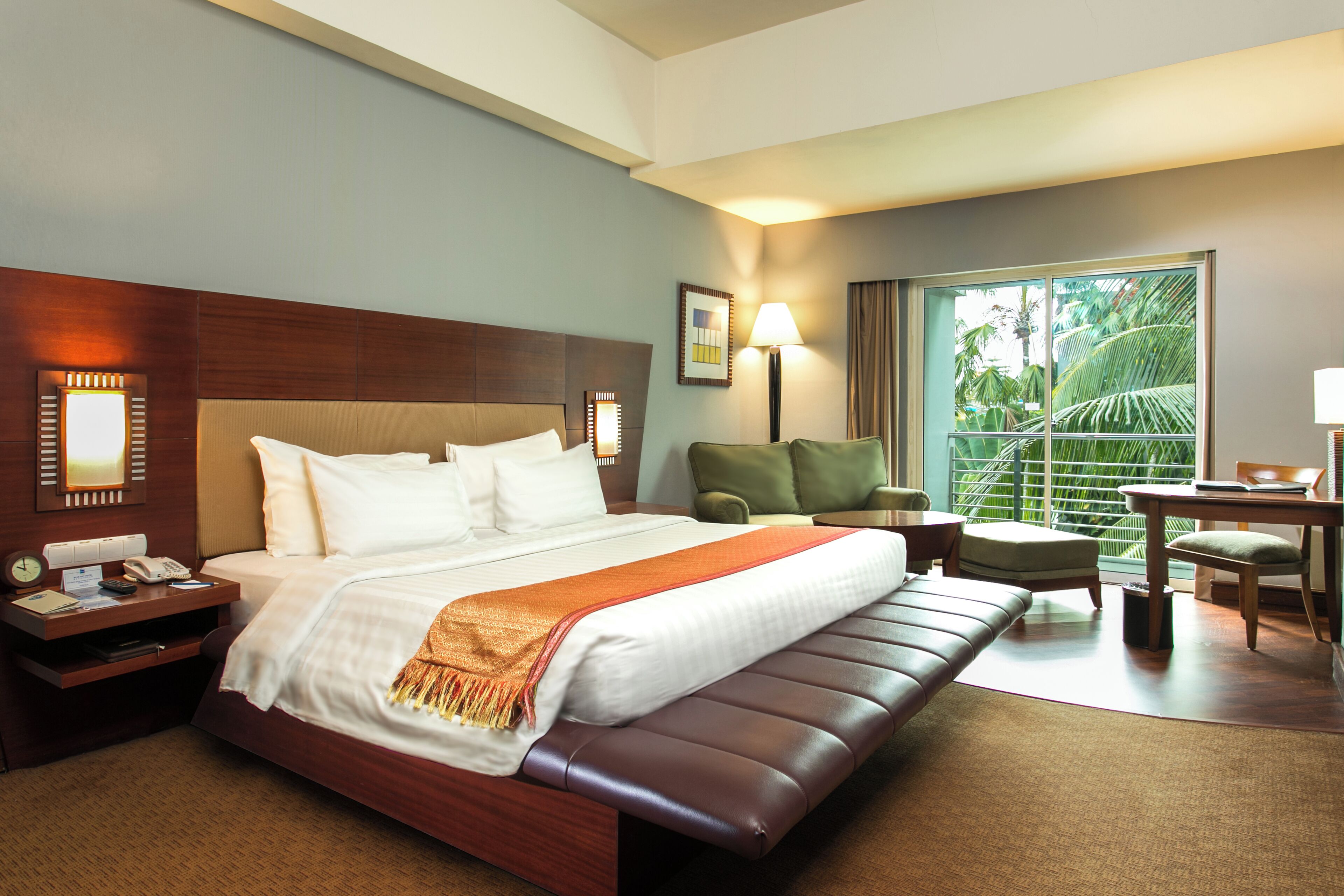 Habitación de hotel con una cama grande, zona de estar con una silla y una mesa, un escritorio con una lámpara, y vistas a zonas verdes por la ventana.
