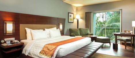Habitación de hotel con una cama grande, zona de estar con una silla y una mesa, un escritorio con una lámpara, y vistas a zonas verdes por la ventana.