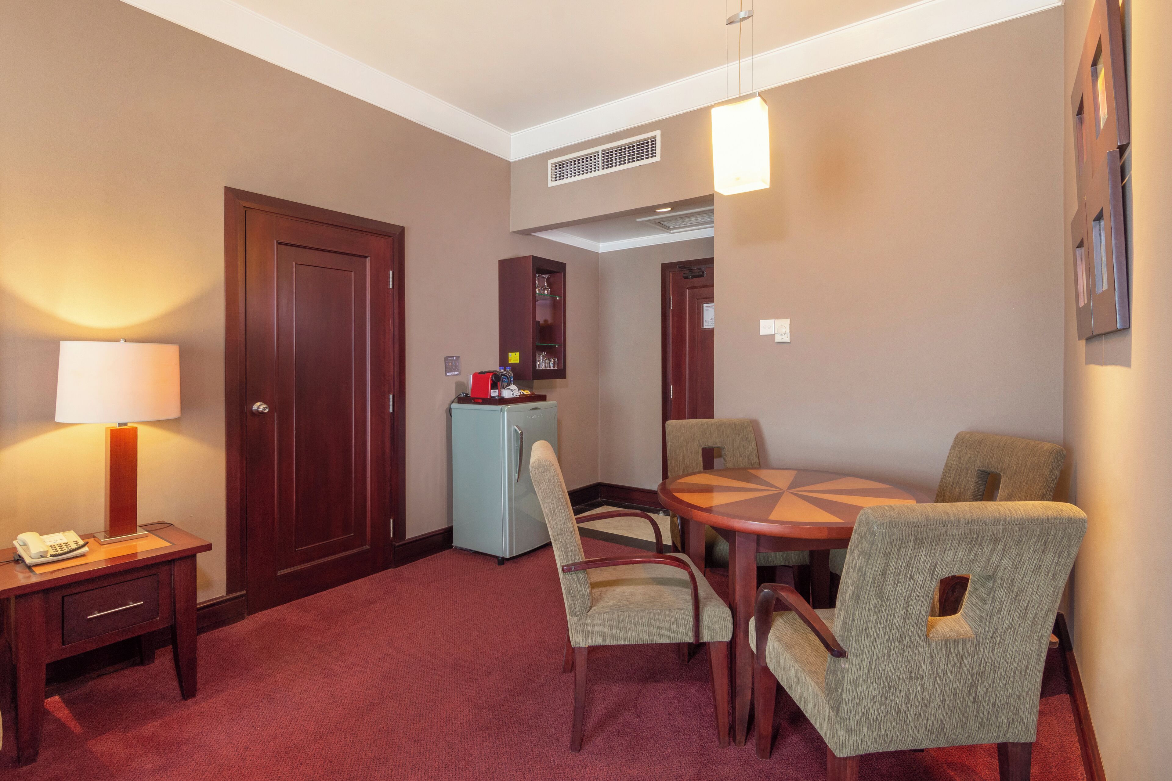 A hotel room with a dining area featuring a round table and four chairs, a small refrigerator, a lamp, and a door.