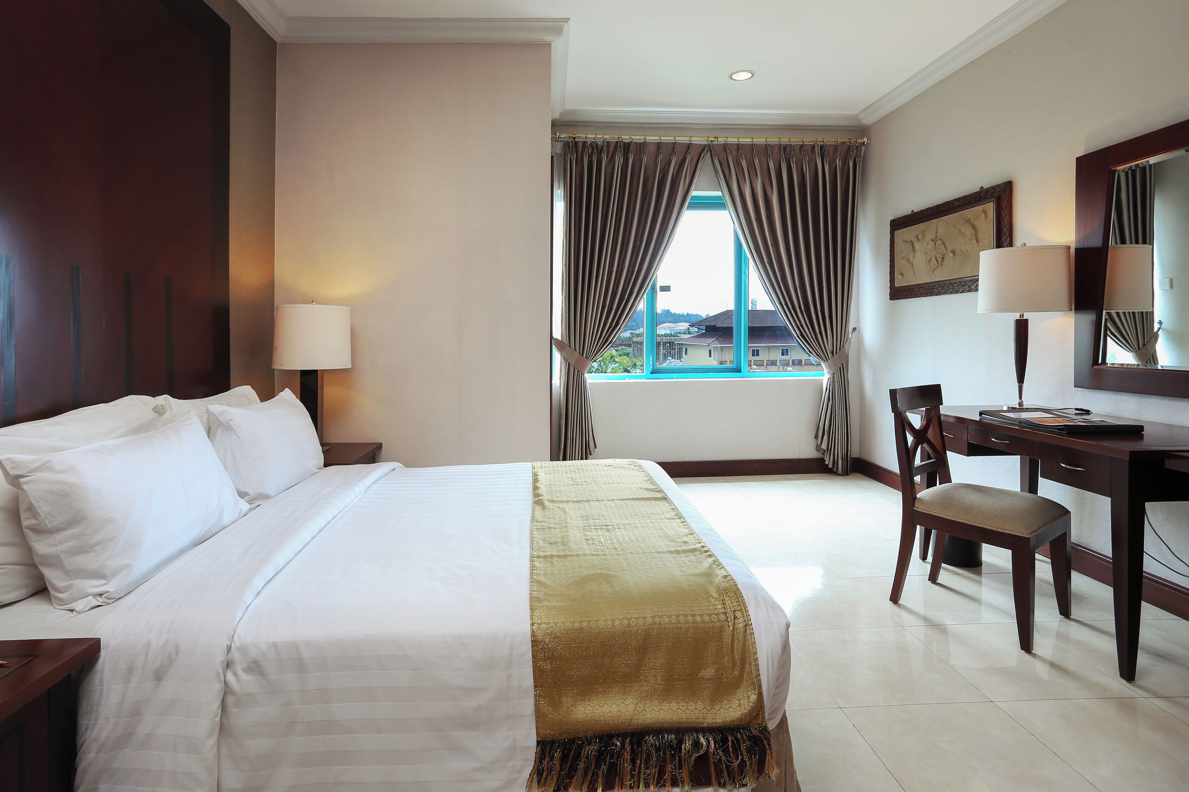 A hotel room with a bed, a desk, a chair, a window with curtains, and a lamp.