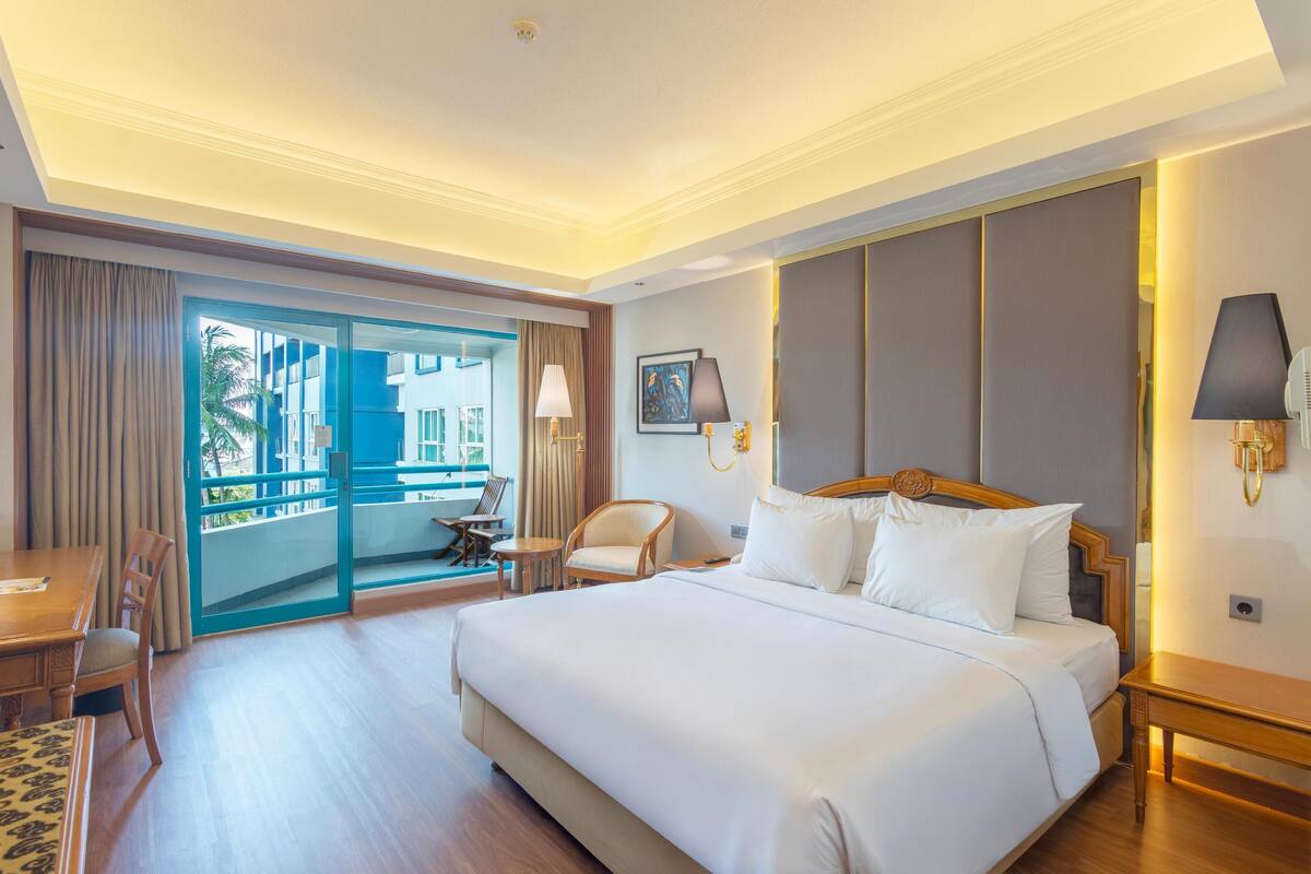 Habitación de hotel con una cama grande, un escritorio, una silla, una mesita y un balcón con vistas a edificios.