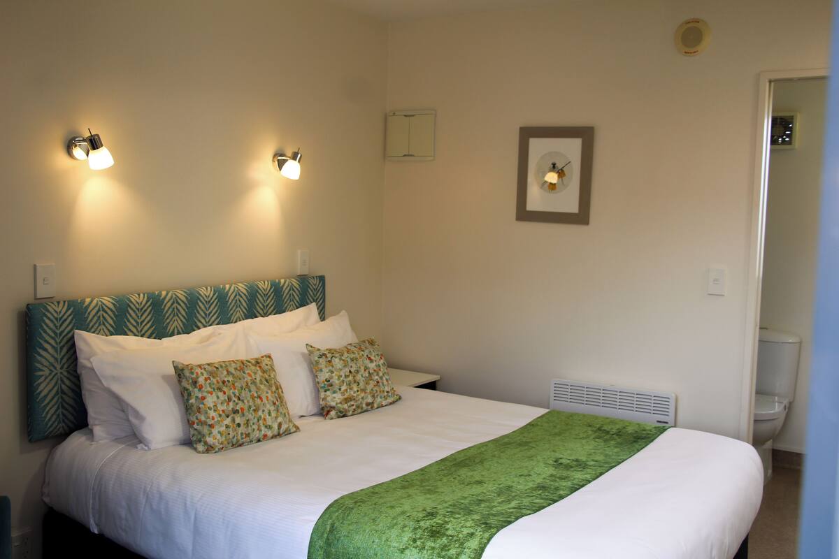 A neatly made bed with a green throw blanket, two wall-mounted reading lights, and a framed picture on the wall.