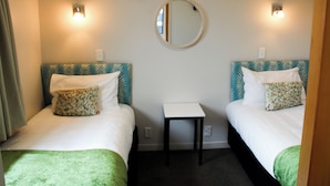 A hotel room with two beds, each with a patterned pillow and a green throw blanket. There is a small table between the beds and a round mirror on the wall. - Bella Vista Motel Mosgiel (Mosgiel)