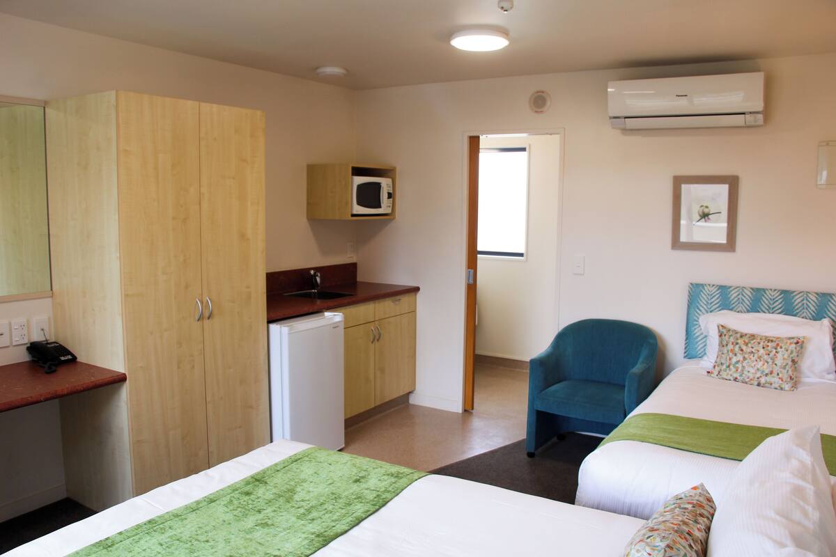 A hotel room with two beds, a desk, a microwave, a chair, and an air conditioning unit.