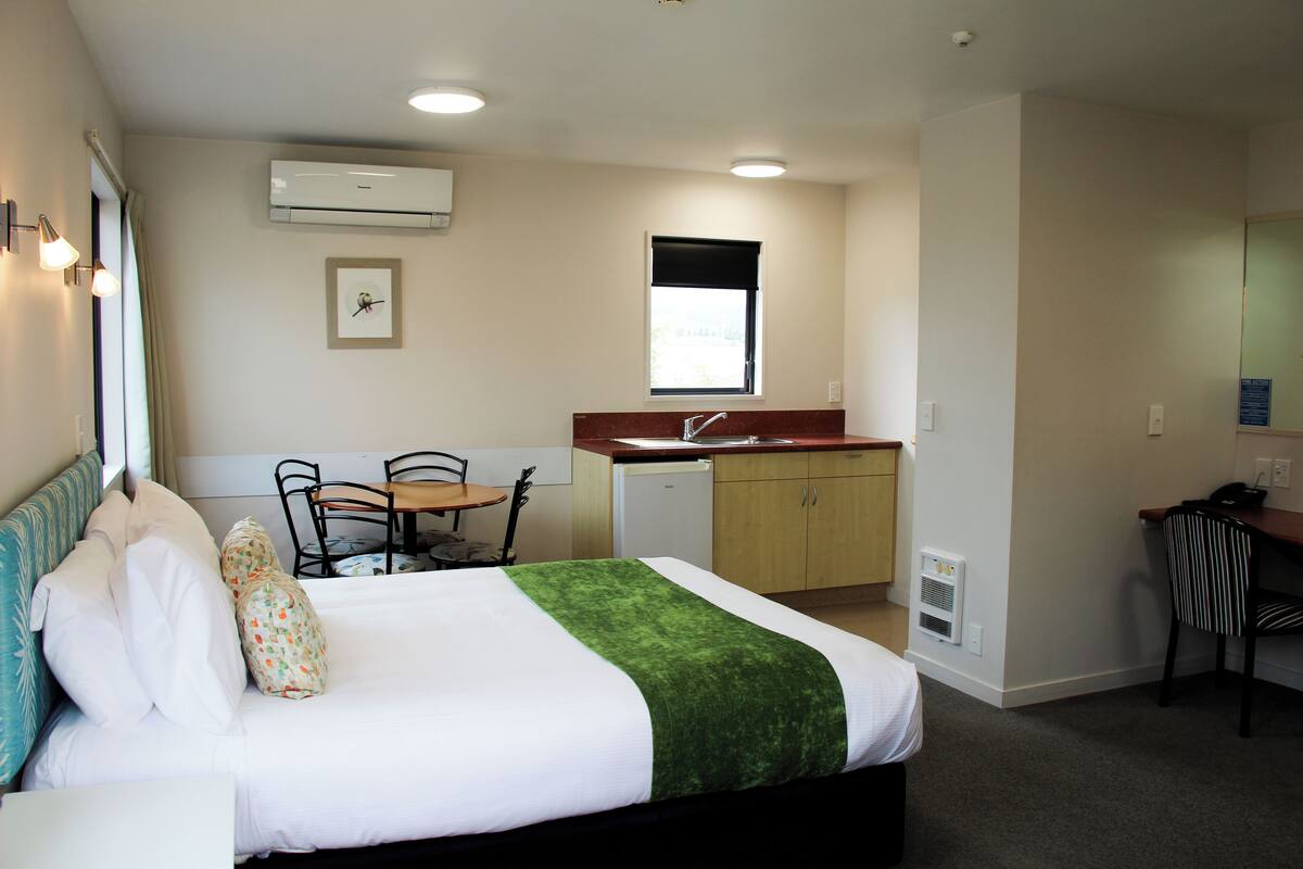 A hotel room with a bed, a small dining table, a chair, a kitchenette, and a window.