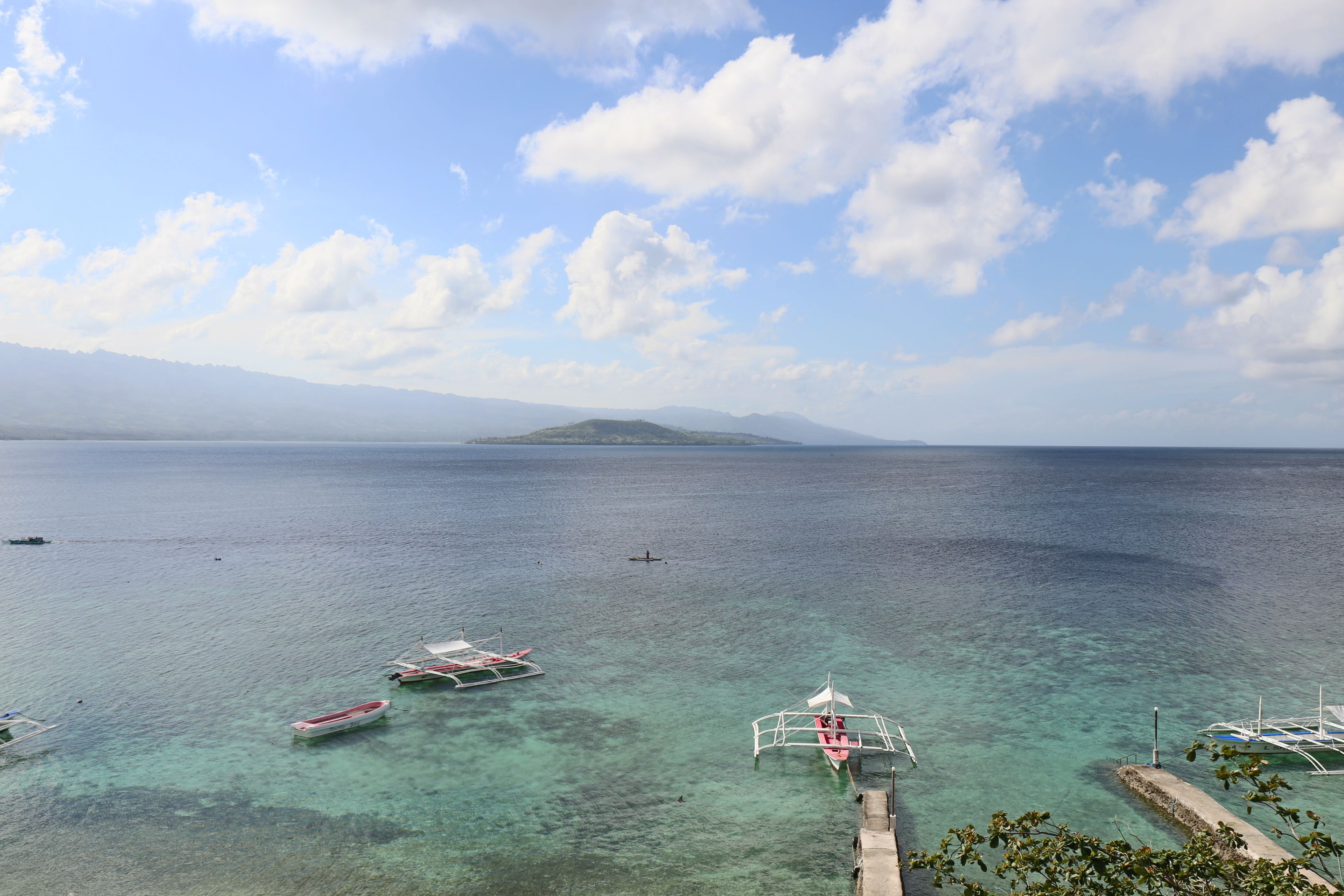 A coastal scene with boats docked at a pier, clear turquoise water, and a distant mountain range under a partly cloudy sky.