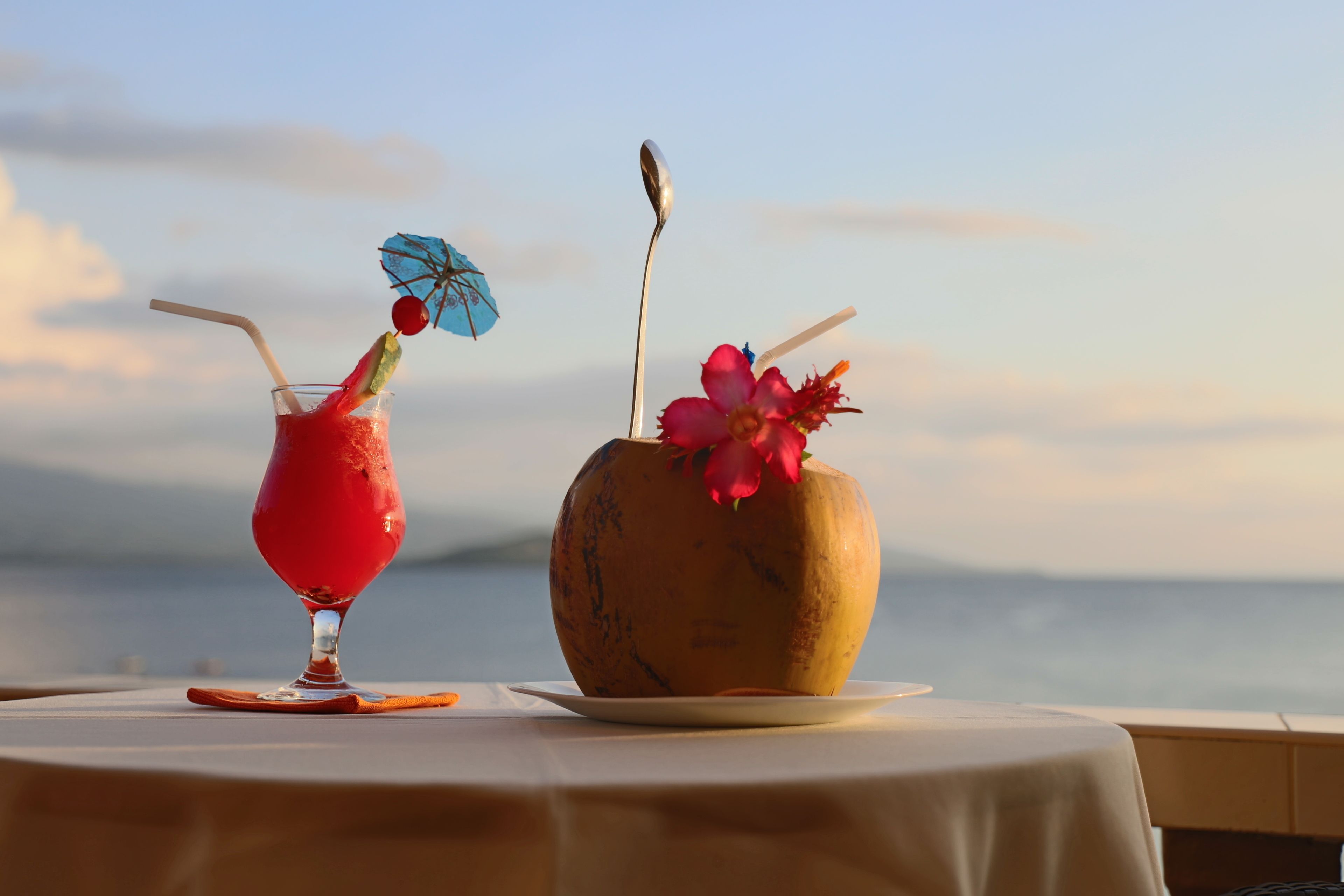 Une table avec une boisson et une noix de coco, le tout posé sur une nappe, avec une vue sur une étendue d’eau.