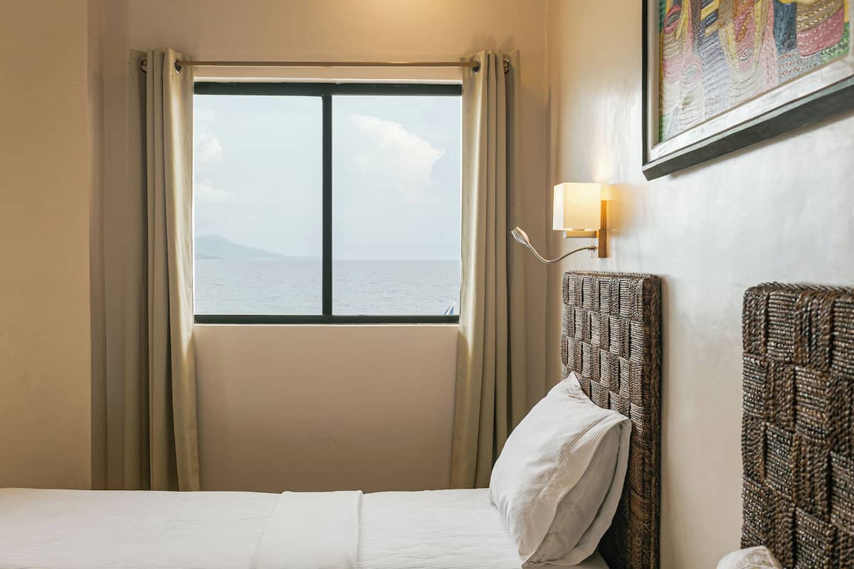 A hotel room with a bed, a window with curtains, a wall-mounted lamp, and a framed picture.