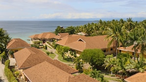 Un resort con varias edificaciones tipo bungalow, cada una con techo de paja, rodeado de exuberante follaje y palmeras. Se observa el océano al fondo, ofreciendo una vista panorámica.