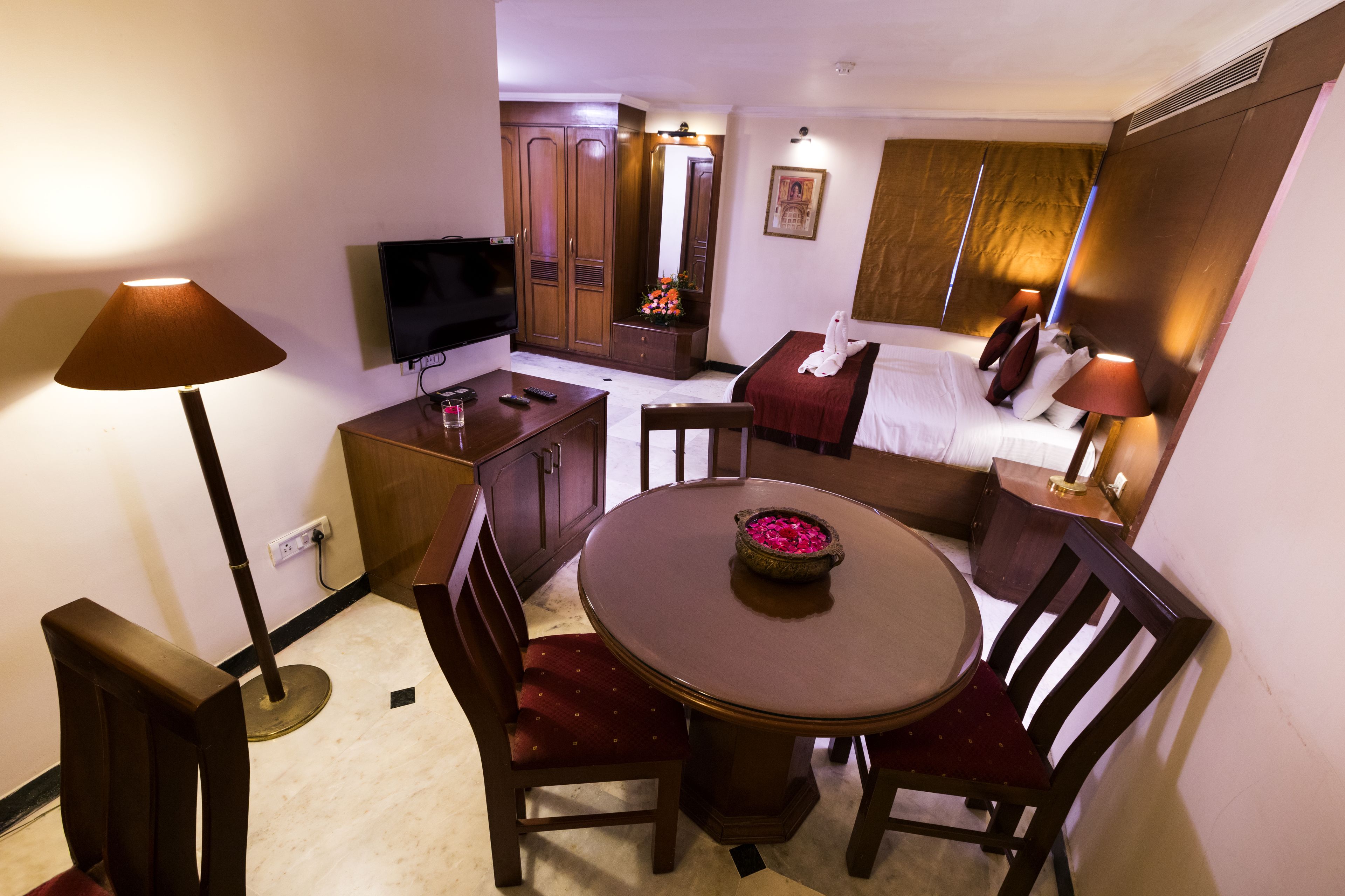 Ein Hotelzimmer mit Esstisch, Stühlen, Fernseher, Kleiderschrank, Bett und Nachttisch mit Lampe.