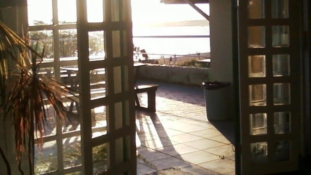 A view through a window with a grid pattern, leading to a patio area with a bench and a view of a body of water. - Pousada Caravelas (Guarapari)