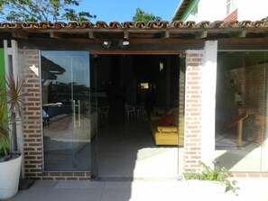 A glass door entrance to a covered outdoor area with a brick wall, a yellow sofa, and a potted plant. - Pousada Caravelas (Guarapari)