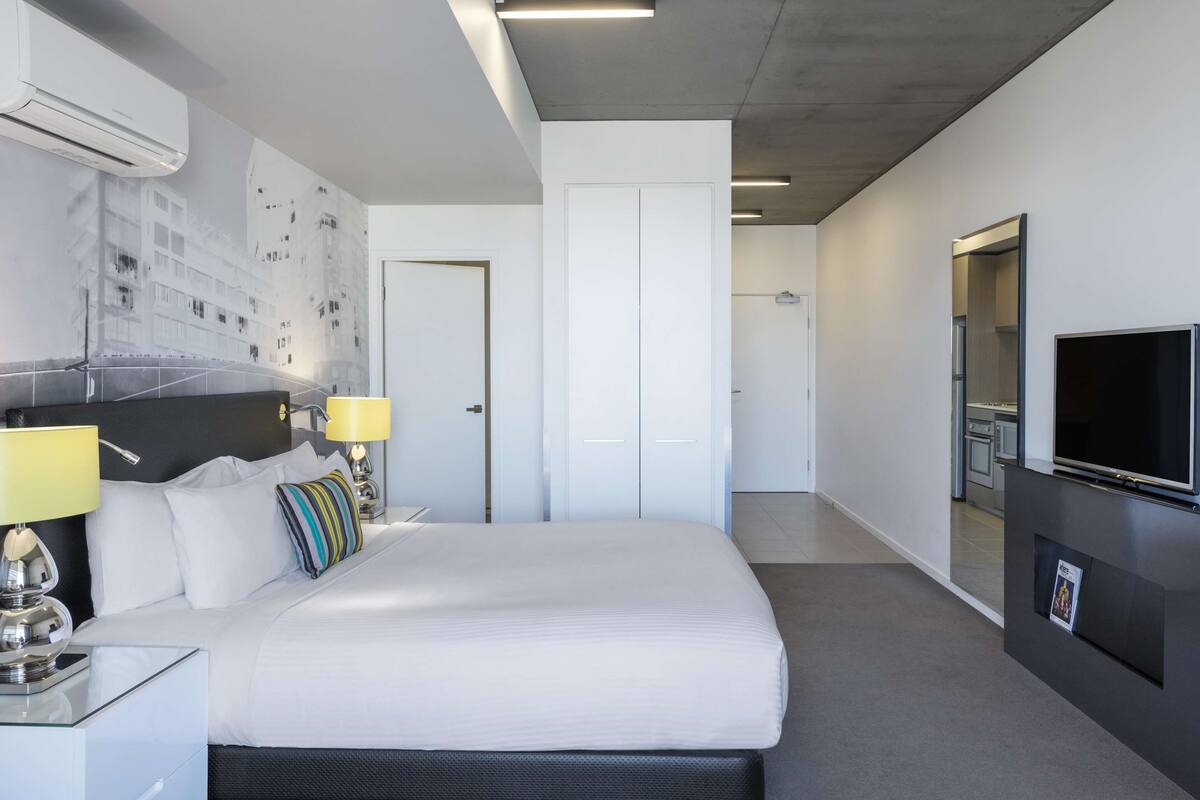 A modern hotel room with a large bed, bedside lamps, a flat-screen TV, and a wall-mounted air conditioner.