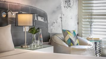 A modern bedroom with a curved headboard, a bedside table with a lamp, a glass vase with a plant, and a chair with a patterned cushion.