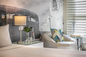 A modern bedroom with a curved headboard, a bedside table with a lamp, a glass vase with a plant, and a chair with a patterned cushion.